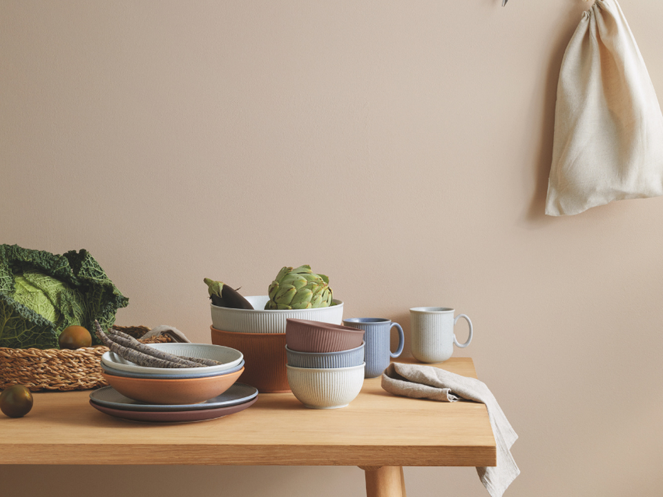 A wooden table with a stack of Thomas crockery in earth tones, cups, kale in a basket and a fabric bag hanging on the wall.