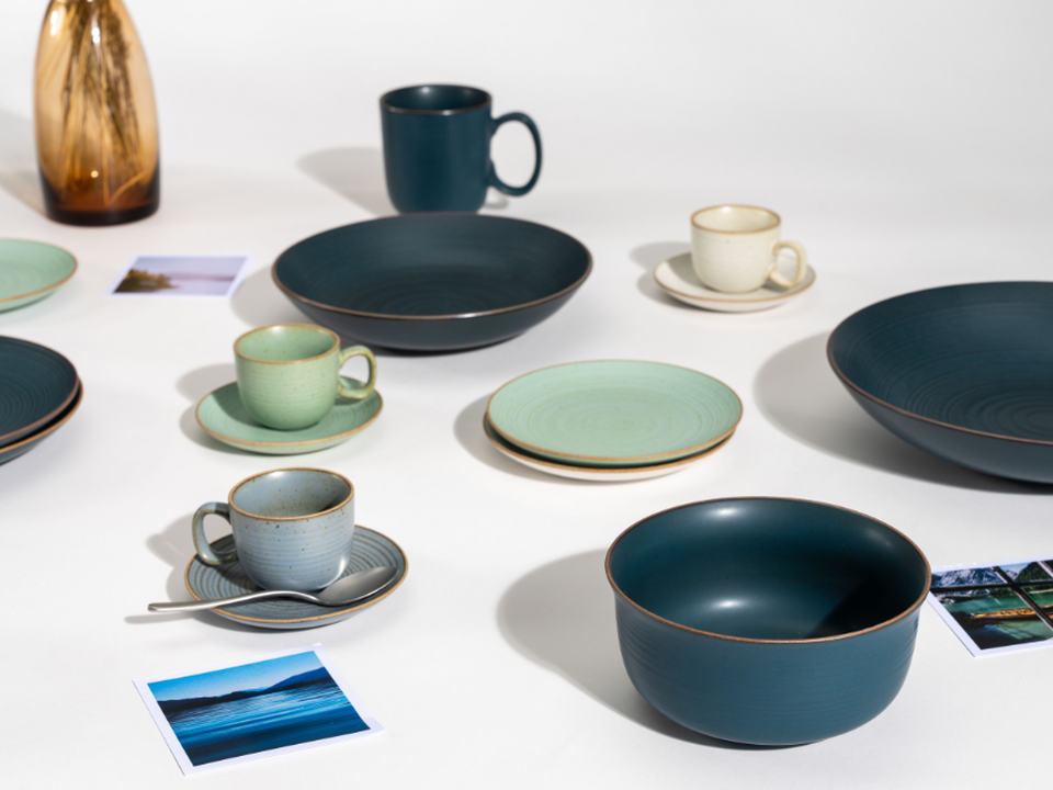 A selection of tableware by Thomas in shades of petrol blue and green: plates, bowls, cups and saucers, and a vase, all set against a white background.