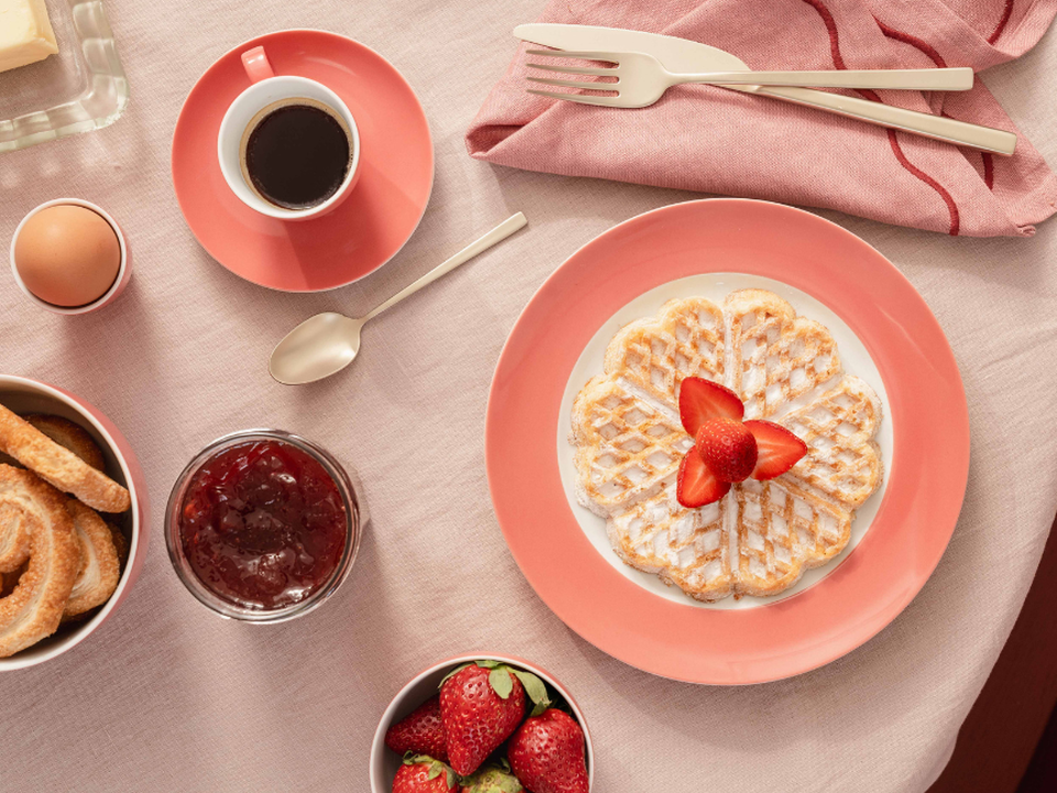 A view from above of the breakfast table, with a waffle and strawberries on a pink plate belonging to Thomas, a coffee cup, jam and toast.