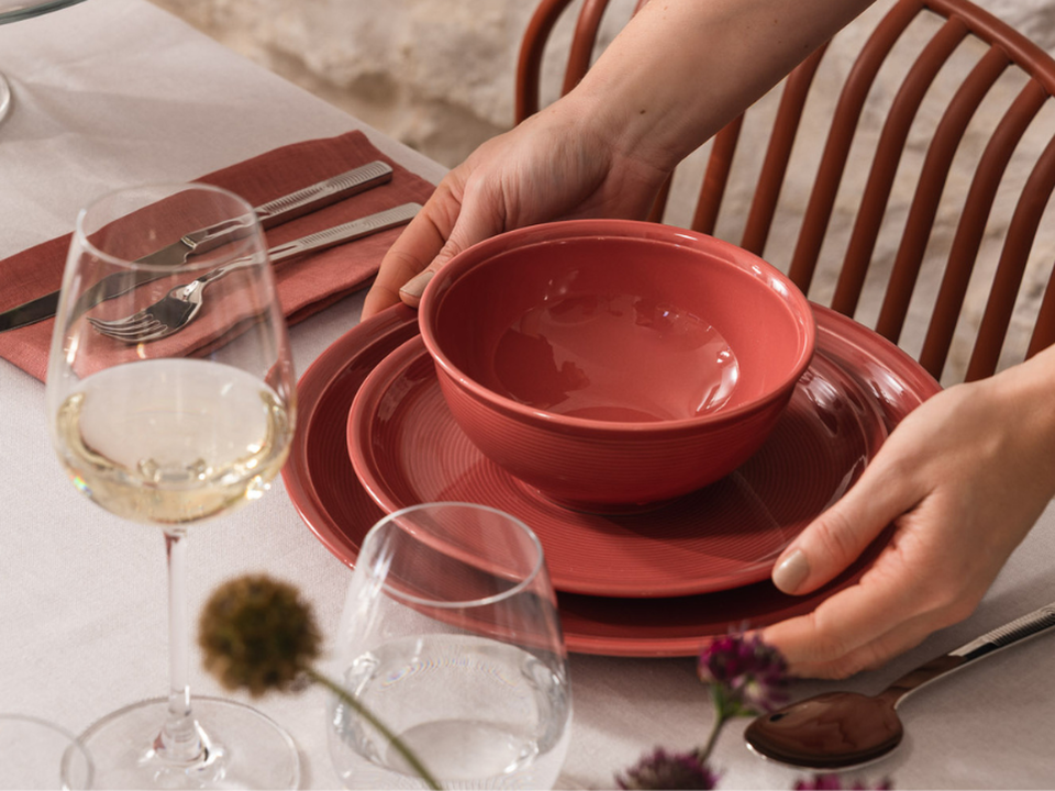 Hands place Thomas&rsquo;s red bowl on a laid table with a red plate, a white wine glass and dried flowers.