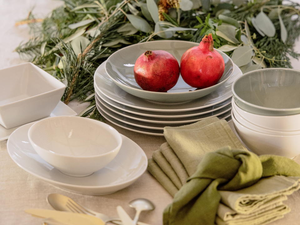 A close-up of a stylishly laid brunch table featuring Thomas tableware in natural cream and green tones: stacked plates and bowls are adorned with two pomegranates. Beside them lie folded cloth napkins in olive green, whilst loosely arranged twigs in the background create a fresh, nature-inspired atmosphere.
