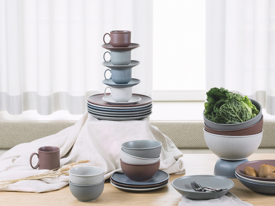 A stack of crockery in pink, grey and blue, with cups on a wooden table in front of a light-coloured curtain.