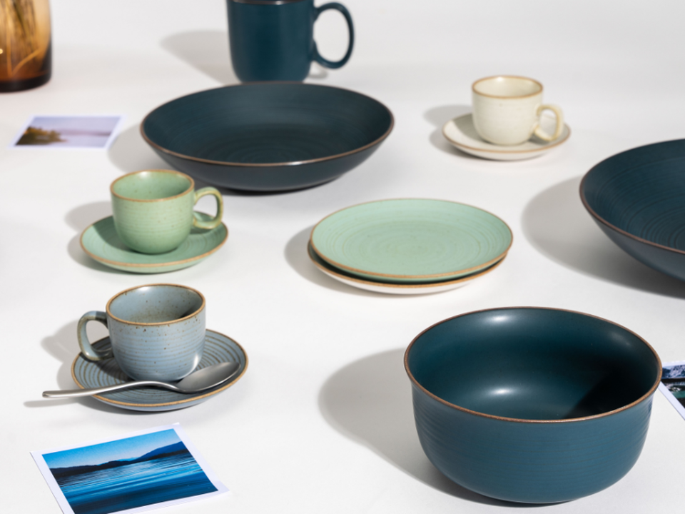 Japandi-style tableware by Thomas, featuring cups, plates and bowls in shades of green, blue and natural tones, arranged on a white table.
