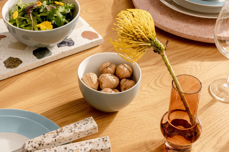 Bright blue bowl filled with walnuts surrounded by a vase with a autumn flower, another sallad bowl, plates placed on a wooden table