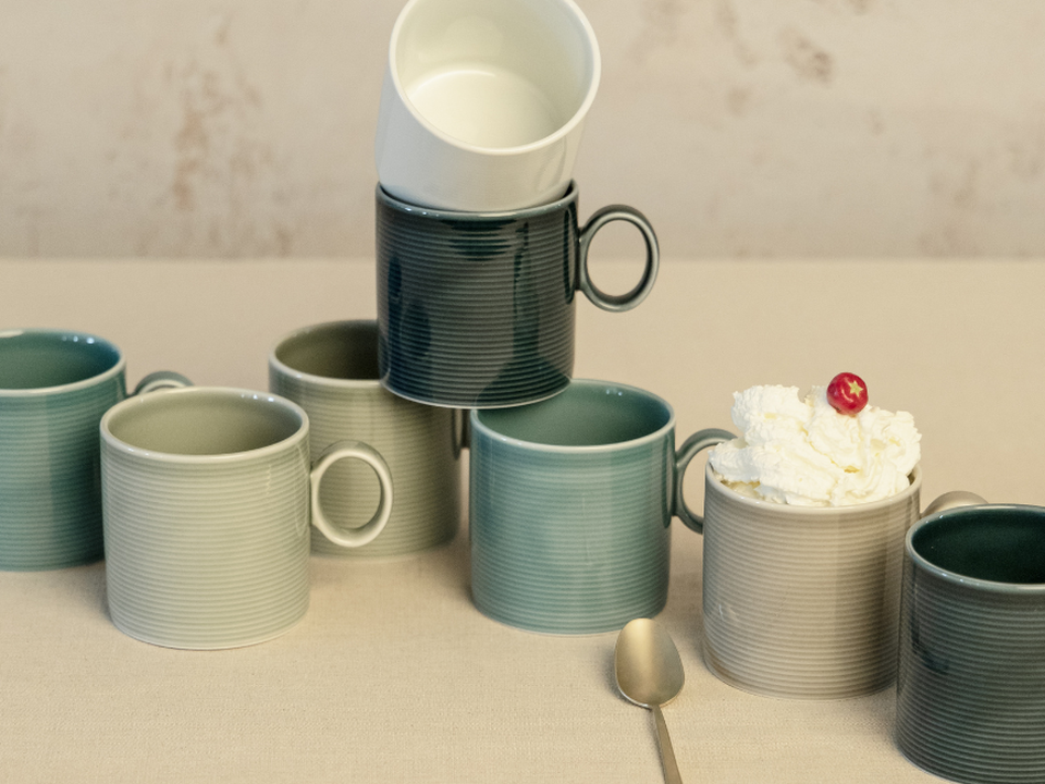 An arrangement of Thomas tableware in soft shades of green and natural tones: several textured cups and mugs are grouped together, some stacked, alongside a bowl containing cream and a cherry.
