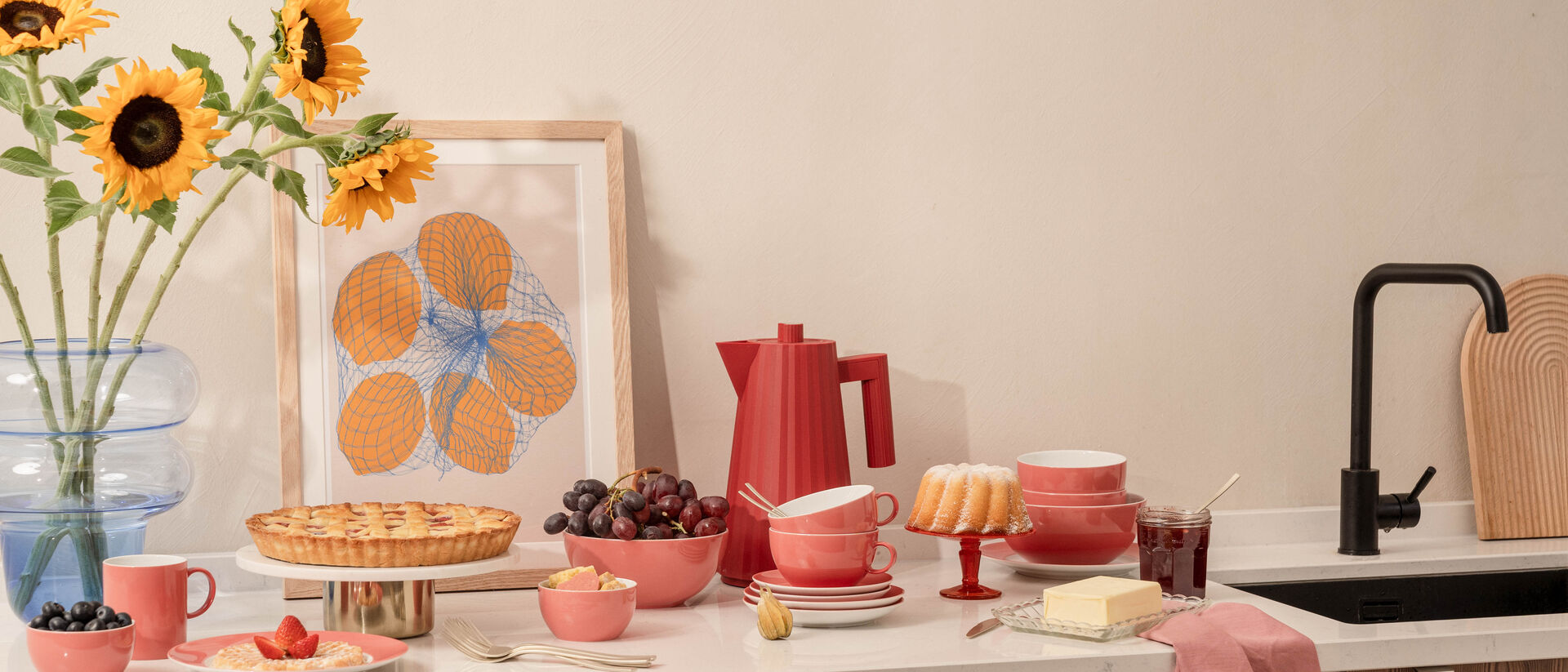 Kitchen worktop with a pink &lsquo;Sunny Day&rsquo; crockery set by Thomas, a coffee pot, sunflowers in a vase, a cake, cherries and a wooden chopping board.
