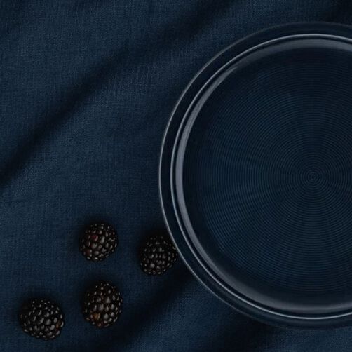 Dark blue plate with blackberries next to it on dark blue background 