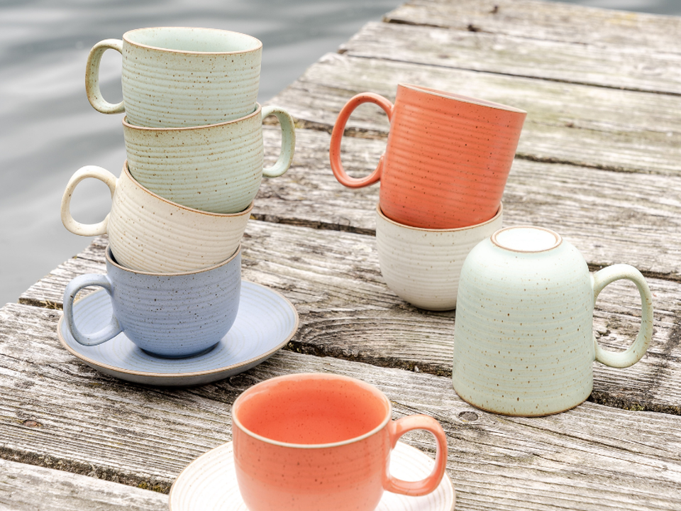 An outdoor brunch scene featuring Thomas tableware in soft pastel and natural tones: several textured cups are arranged on a wooden table, some stacked and others placed on saucers.
