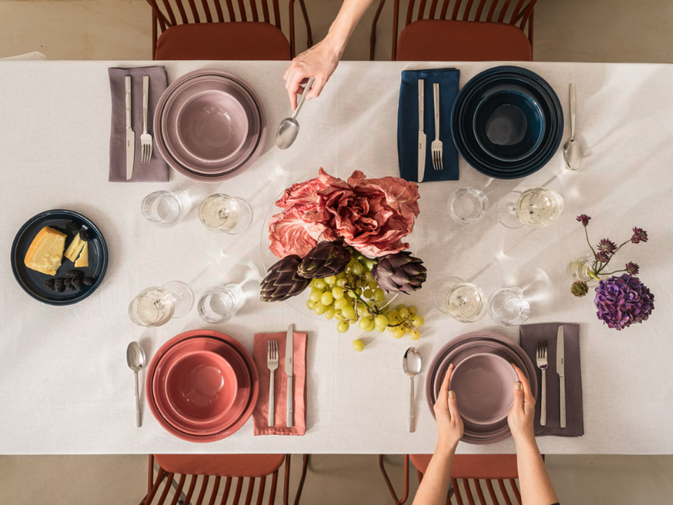 A photograph taken from above of a festively laid table with colourful crockery, glasses, a cheese platter, grapes and flowers; hands are setting the table.