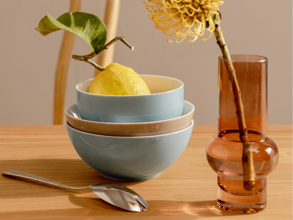 A stack of light blue bowls by Thomas, with a lemon on a wooden table; next to them are spoons and a vase containing a yellow dried flower.