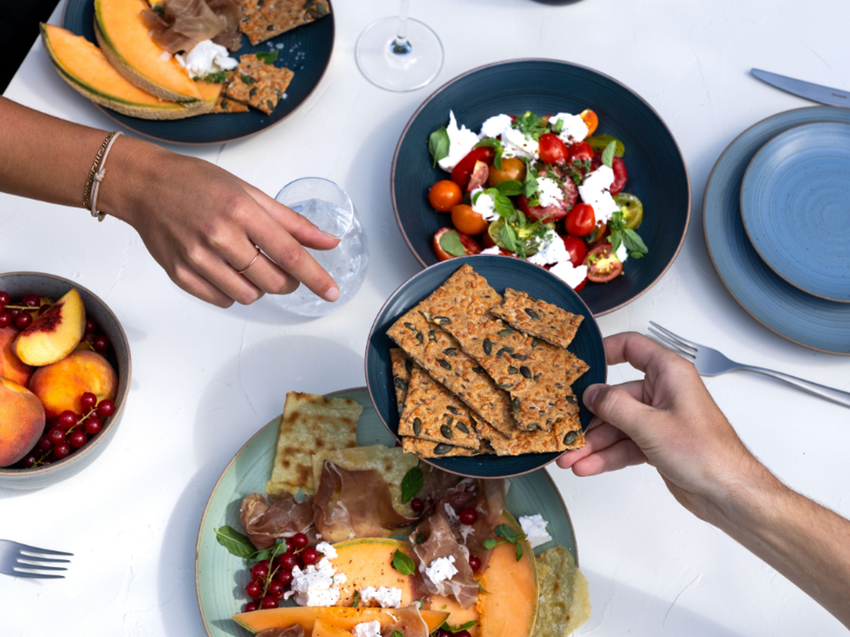 A table photographed from above, with salad, bread, melon and fruit on dark plates; hands as people eat together.