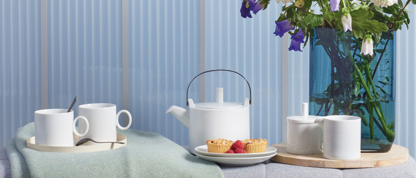 Wei&szlig;e Tassen, Teekanne und Milchk&auml;nnchen mit Zuckerdose auf einer Bank mit Blumenvase im HIntergrund