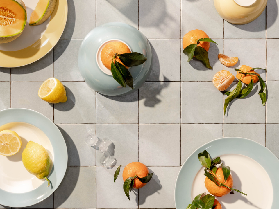 A table photographed from above, featuring citrus fruits and light blue and white plates on a tiled surface.