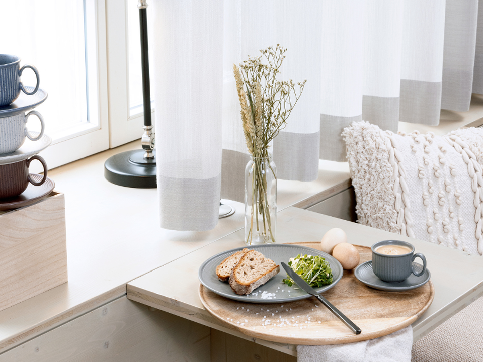 A breakfast table with bread, an egg, a coffee cup and a vase of grasses in front of a light-coloured curtain, with a stack of cups in the background.