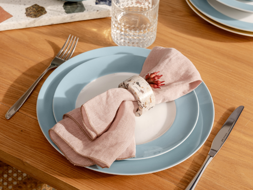 A close-up of a set brunch table featuring Thomas tableware in a delicate light blue: one plate is set with a folded fabric napkin in a shade of ros&eacute; and a decorative napkin ring. Cutlery and a water glass complete the minimalist, warm table setting on a wooden surface.