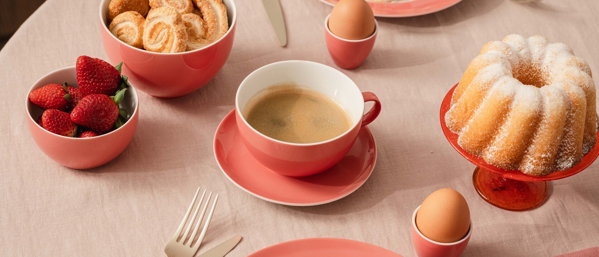 A brunch scene featuring colour-coordinated Thomas tableware in warm shades of ros&eacute; and coral: on a laid table sit a cup of coffee, soft-boiled eggs in egg cups, fresh strawberries, pastries and a Gugelhupf on a cake stand.