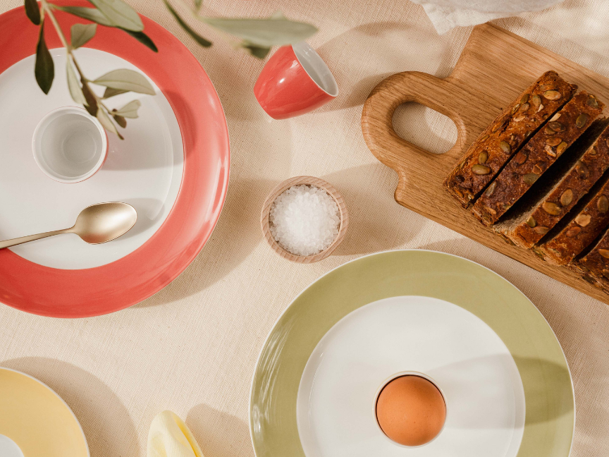 Draufsicht auf eine stilvoll gedeckte Brunchszene mit Thomas-Geschirr: Farbige Teller in Koralle und Gr&uuml;n rahmen ein schlichtes wei&szlig;es Zentrum mit einem Ei im Eierbecher. Daneben liegen geschnittenes Brot auf einem Holzbrett, ein kleines Sch&auml;lchen mit Salz sowie minimalistisch arrangiertes Besteck und Servietten in harmonischen Naturt&ouml;nen.