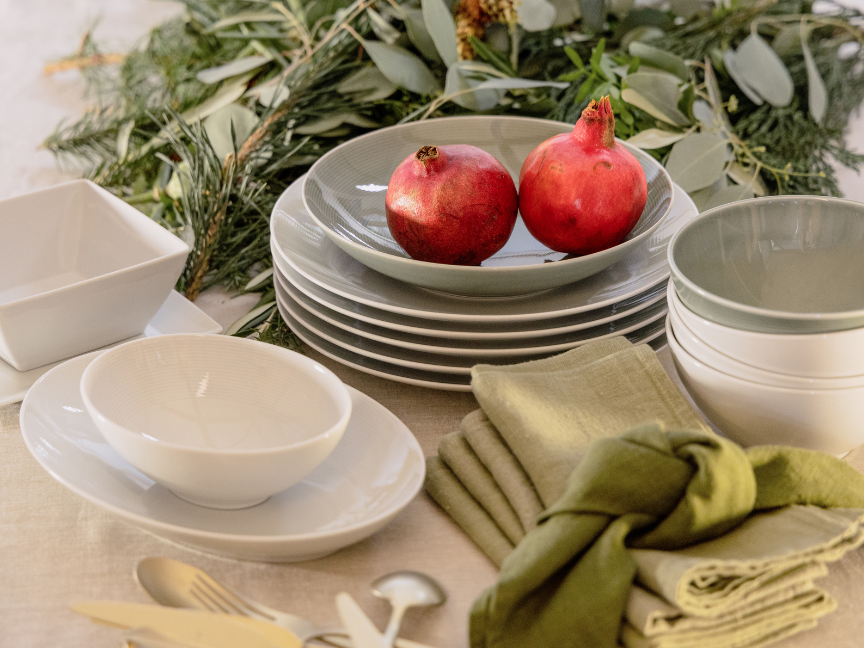 Detailaufnahme eines stilvoll gedeckten Brunch-Tisches mit Thomas-Geschirr in nat&uuml;rlichen Creme- und Gr&uuml;nt&ouml;nen: Gestapelte Teller und Schalen werden von zwei Granat&auml;pfeln dekorativ gekr&ouml;nt. Daneben liegen gefaltete Stoffservietten in Olivgr&uuml;n, w&auml;hrend locker arrangierte Zweige im Hintergrund eine frische, naturinspirierte Atmosph&auml;re schaffen.