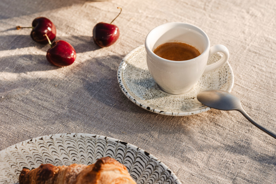 Thomas Sandora creamwhite espresso cup on espresso saucer in lightbeige on beige tablecloth with cherries laying in the background