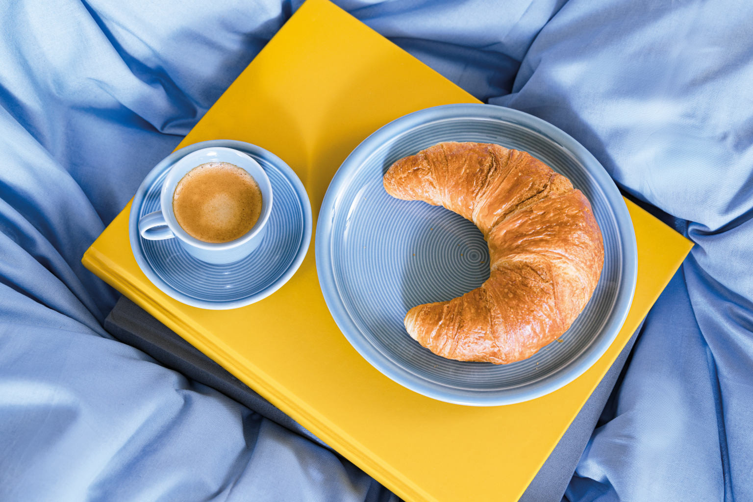 Thomas Trend Arctic Blue Teller mit Croissant und Kaffeetasse auf gelben Buch auf blauer Bettdecke