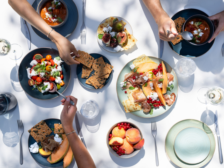 Draufsicht auf eine lebendige Brunchszene mit Thomas-Geschirr: Mehrere Personen greifen gleichzeitig nach Speisen auf modernem Geschirr in verschiedenen Farben. Serviert werden unter anderem frischer Salat, Brot, Obst und ein gro&szlig;es Gericht in der Mitte.