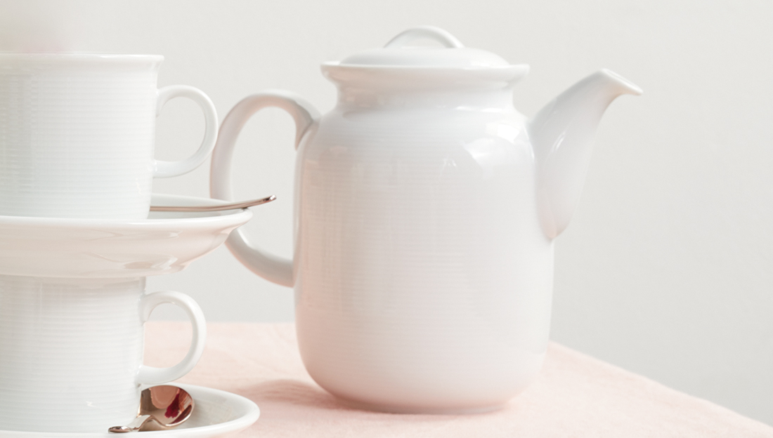 Teekanne auf Tisch neben zwei Kaffeetassen mit Untersetzer und Löffel